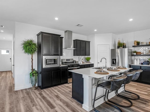 kitchen featuring an island with sink, backsplash, wall chimney exhaust hood, stainless steel appliances, and sink