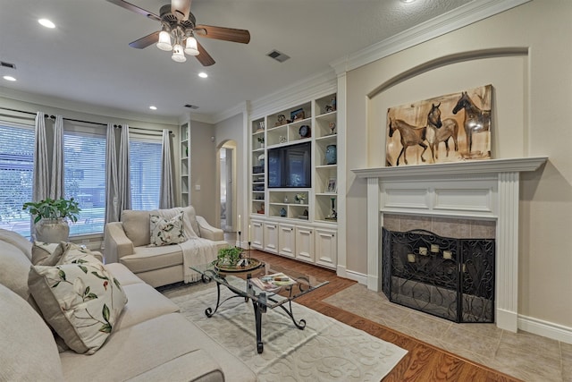 living room featuring built in shelves, ceiling fan, light wood-type flooring, a fireplace, and ornamental molding