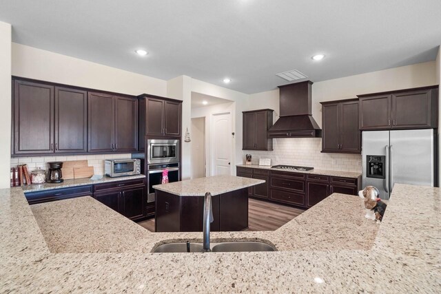 kitchen featuring light stone counters, dark brown cabinets, a sink, stainless steel appliances, and premium range hood