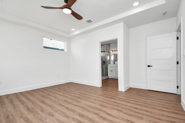 unfurnished bedroom featuring ensuite bathroom, ceiling fan, light wood-type flooring, and a tray ceiling