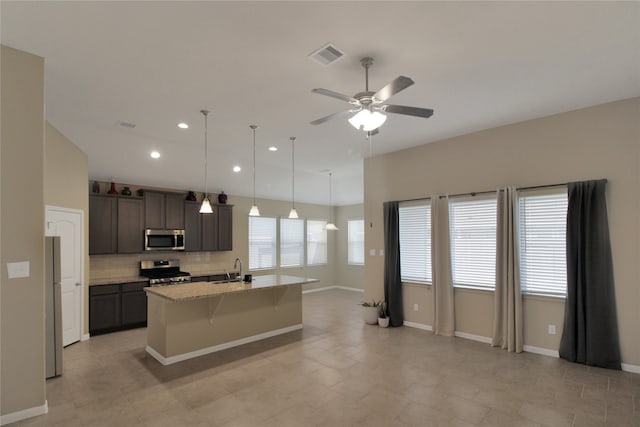 kitchen featuring stainless steel appliances, dark brown cabinetry, light stone counters, decorative backsplash, and a kitchen island with sink