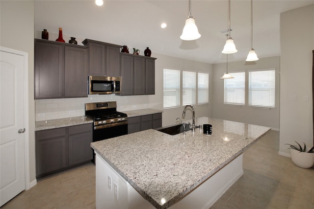 kitchen featuring a kitchen island with sink, stainless steel appliances, sink, decorative light fixtures, and light stone counters