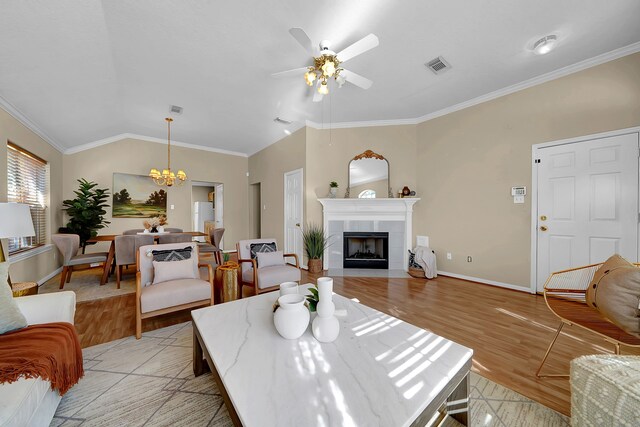 living room with ceiling fan with notable chandelier, a tiled fireplace, vaulted ceiling, crown molding, and light wood-type flooring