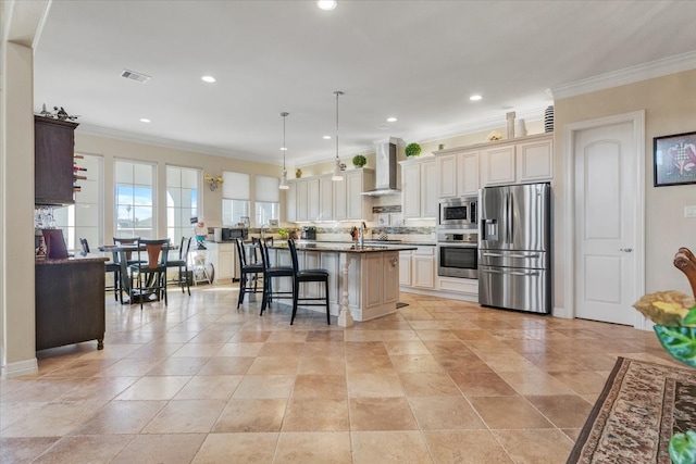 kitchen featuring an island with sink, a breakfast bar, wall chimney exhaust hood, stainless steel appliances, and cream cabinets