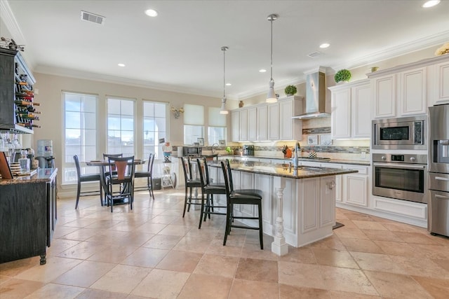 kitchen featuring dark stone counters, an island with sink, wall chimney range hood, stainless steel appliances, and ornamental molding