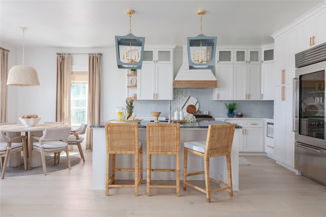 kitchen featuring white cabinetry, light hardwood / wood-style flooring, backsplash, decorative light fixtures, and premium range hood