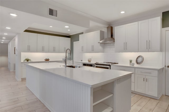 kitchen with wall chimney range hood, an island with sink, and white cabinets