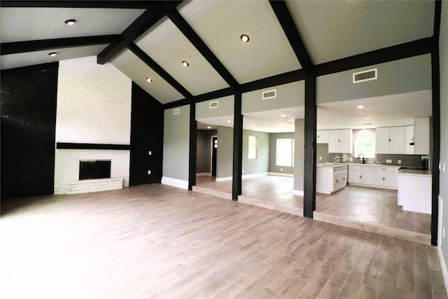 unfurnished living room featuring a brick fireplace, beam ceiling, light hardwood / wood-style flooring, sink, and high vaulted ceiling