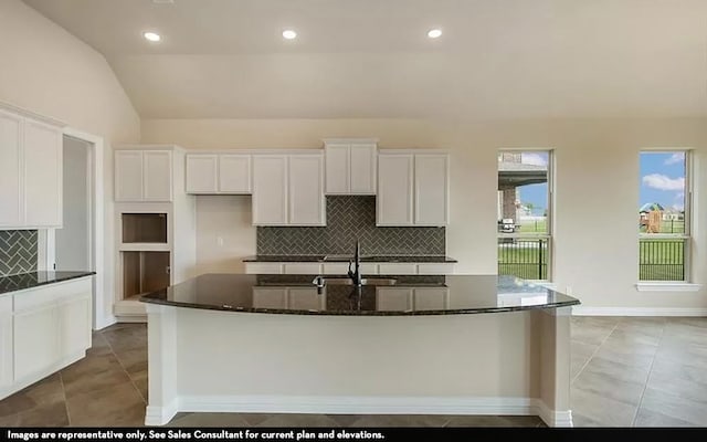 kitchen featuring dark stone counters, a kitchen island with sink, white cabinets, lofted ceiling, and decorative backsplash