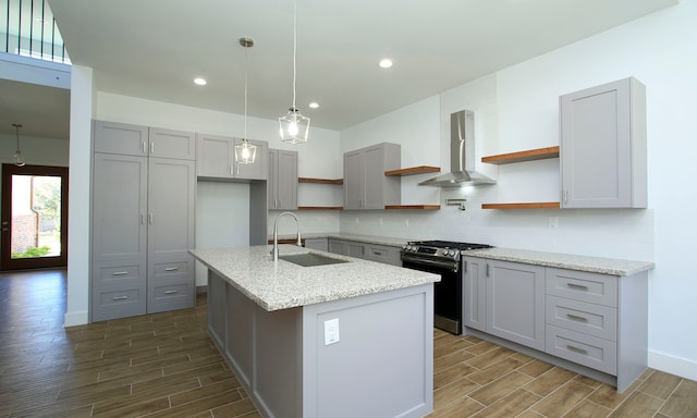 kitchen with light stone countertops, a kitchen island with sink, sink, wall chimney range hood, and stainless steel range oven