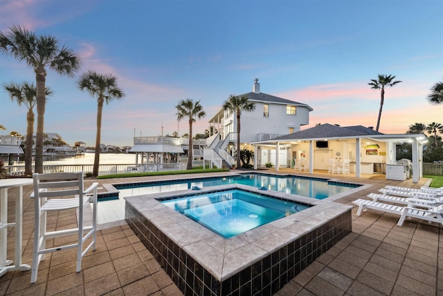 pool at dusk featuring a water view, an in ground hot tub, and a patio