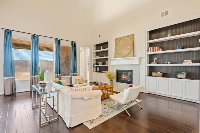 living room featuring a tile fireplace, built in shelves, a towering ceiling, and dark hardwood / wood-style floors