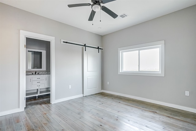 unfurnished bedroom with ceiling fan, light hardwood / wood-style flooring, ensuite bath, and a barn door