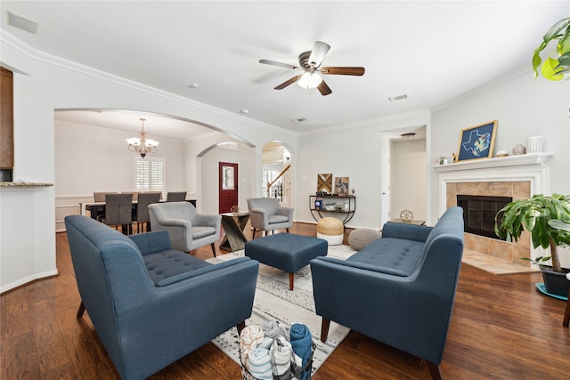 living room featuring ornamental molding, ceiling fan with notable chandelier, a tiled fireplace, and dark wood-type flooring