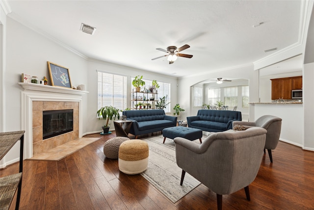 living room featuring ceiling fan, ornamental molding, a tiled fireplace, and dark hardwood / wood-style flooring