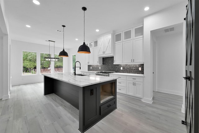 kitchen featuring backsplash, white cabinets, a center island with sink, sink, and light stone countertops