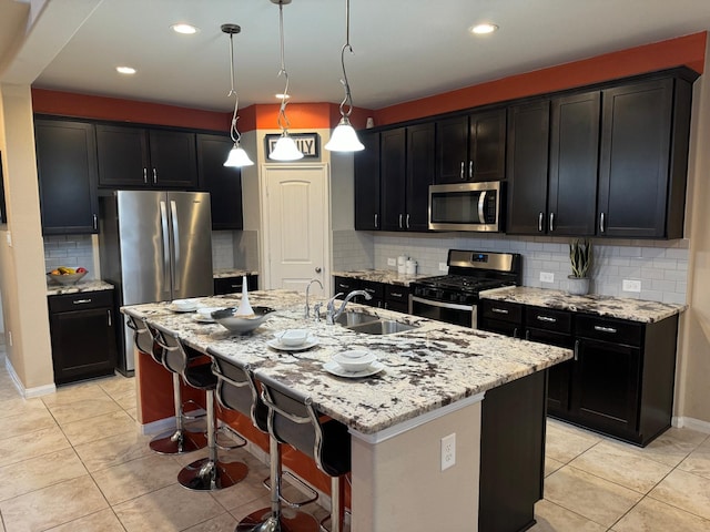 kitchen featuring a kitchen island with sink, hanging light fixtures, stainless steel appliances, light stone counters, and decorative backsplash