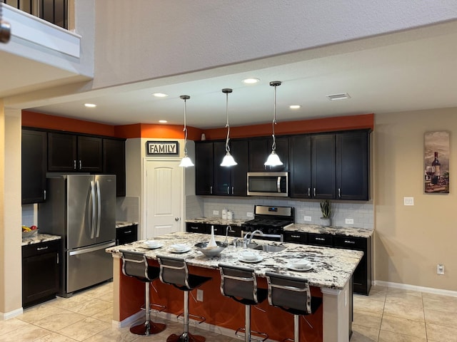 kitchen featuring hanging light fixtures, stainless steel appliances, light stone counters, an island with sink, and a kitchen bar