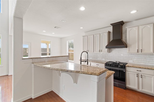 kitchen featuring light stone counters, stainless steel range with gas stovetop, wall chimney range hood, white cabinetry, and light wood-type flooring