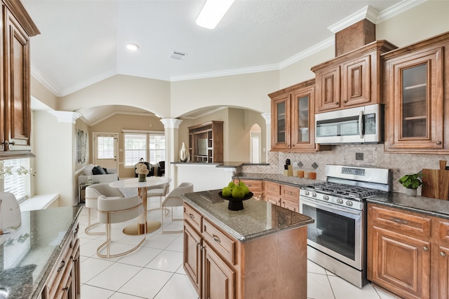 kitchen with stainless steel appliances, decorative columns, dark stone counters, crown molding, and vaulted ceiling