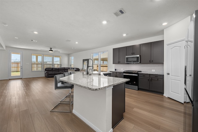 kitchen with light hardwood / wood-style floors, an island with sink, light stone countertops, stainless steel appliances, and ceiling fan