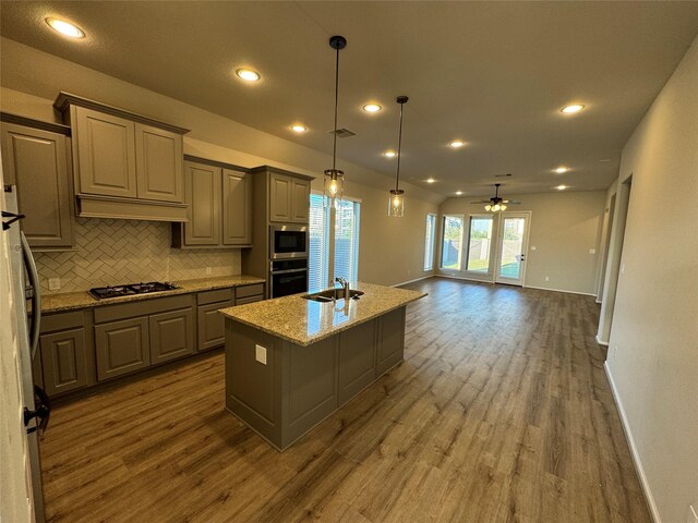 kitchen with appliances with stainless steel finishes, light stone counters, dark hardwood / wood-style flooring, ceiling fan, and a center island with sink