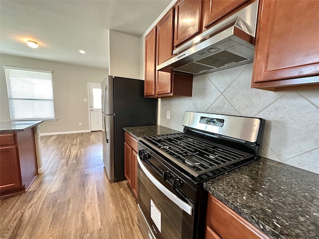 kitchen with appliances with stainless steel finishes, decorative backsplash, exhaust hood, light wood-type flooring, and dark stone counters