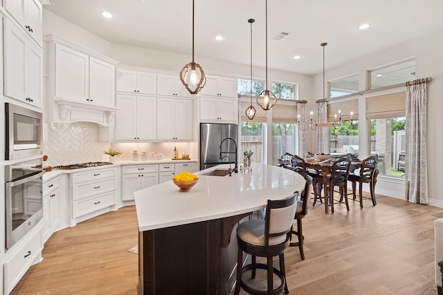 kitchen featuring hanging light fixtures, a kitchen island with sink, white cabinetry, stainless steel appliances, and light wood-type flooring