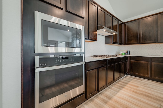 kitchen featuring light wood-type flooring, vaulted ceiling, decorative backsplash, appliances with stainless steel finishes, and dark brown cabinetry