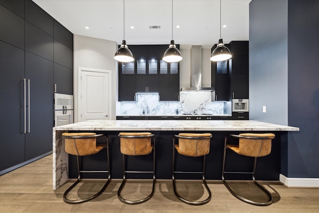 kitchen with wall chimney range hood, decorative light fixtures, tasteful backsplash, a large island, and light hardwood / wood-style floors