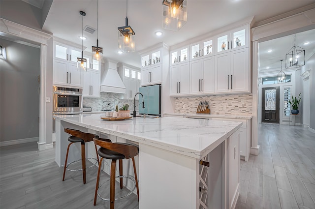 kitchen featuring pendant lighting, sink, a kitchen island with sink, custom range hood, and white cabinets