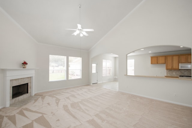 unfurnished living room featuring ceiling fan, high vaulted ceiling, a tile fireplace, light carpet, and crown molding