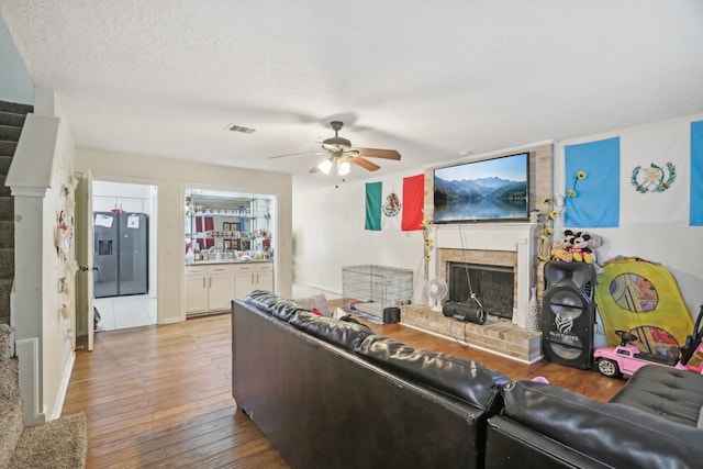 living room featuring light wood-type flooring, a textured ceiling, ceiling fan, and a brick fireplace