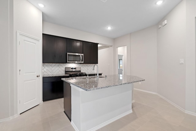 kitchen featuring light stone counters, a kitchen island with sink, a sink, appliances with stainless steel finishes, and backsplash