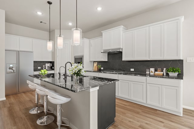 kitchen with dark stone countertops, white cabinetry, hanging light fixtures, appliances with stainless steel finishes, and an island with sink