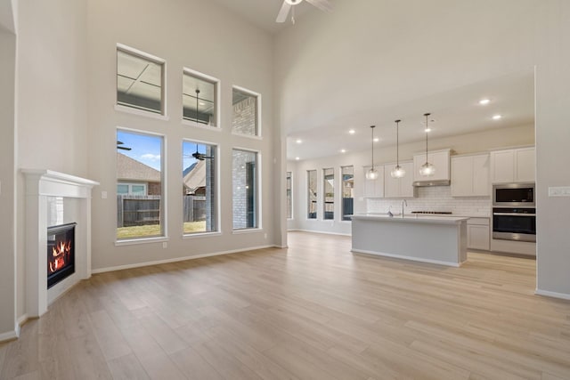 unfurnished living room featuring ceiling fan, a towering ceiling, baseboards, light wood finished floors, and a glass covered fireplace