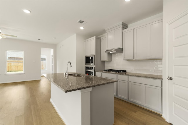 kitchen featuring sink, gray cabinetry, appliances with stainless steel finishes, an island with sink, and dark stone counters