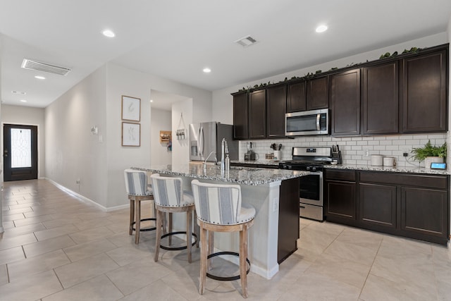 kitchen featuring a center island with sink, decorative backsplash, appliances with stainless steel finishes, dark brown cabinets, and light stone counters