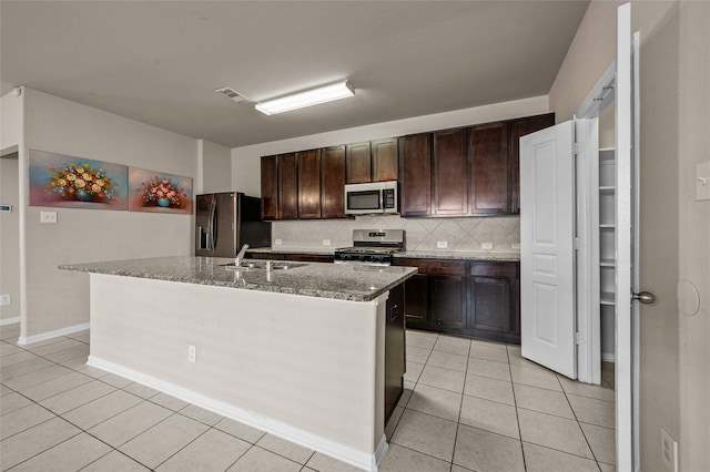 kitchen with light stone countertops, appliances with stainless steel finishes, dark brown cabinetry, backsplash, and a center island with sink