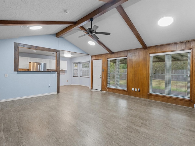 unfurnished living room featuring wood walls, light hardwood / wood-style flooring, vaulted ceiling with beams, ceiling fan, and a textured ceiling