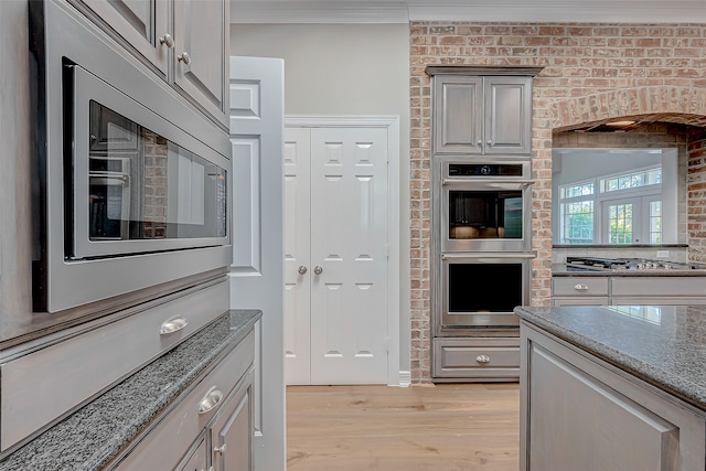 kitchen with gray cabinetry, brick wall, dark stone counters, appliances with stainless steel finishes, and light wood-type flooring