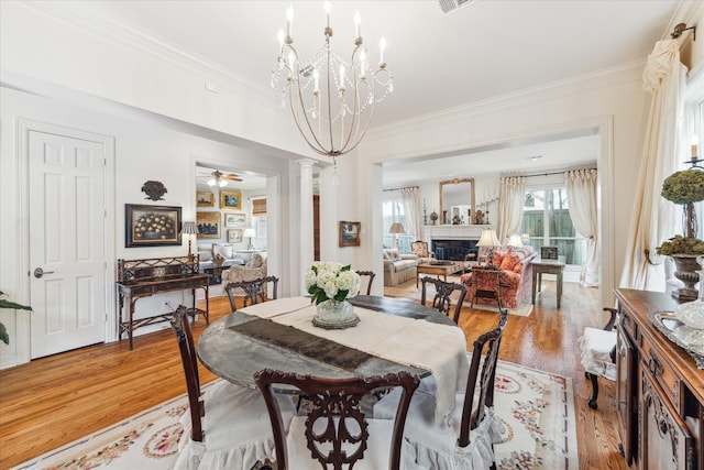 dining room featuring a fireplace, decorative columns, light wood-style flooring, ornamental molding, and ceiling fan with notable chandelier