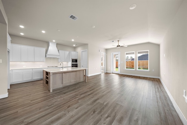 kitchen featuring white cabinets, wood-type flooring, a center island with sink, custom range hood, and stainless steel microwave