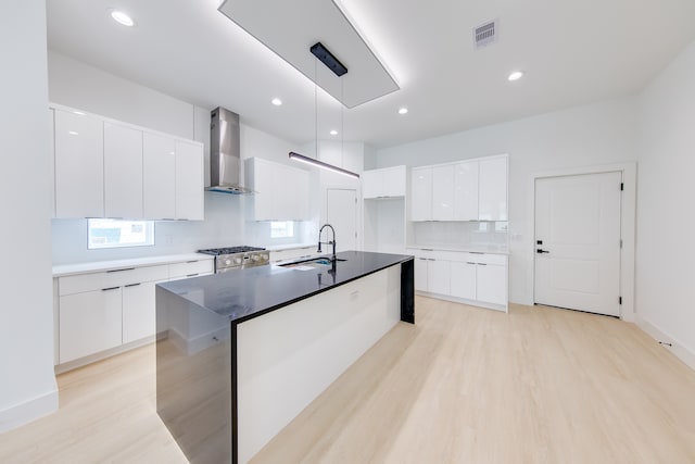 kitchen featuring hanging light fixtures, white cabinetry, a center island with sink, sink, and wall chimney range hood