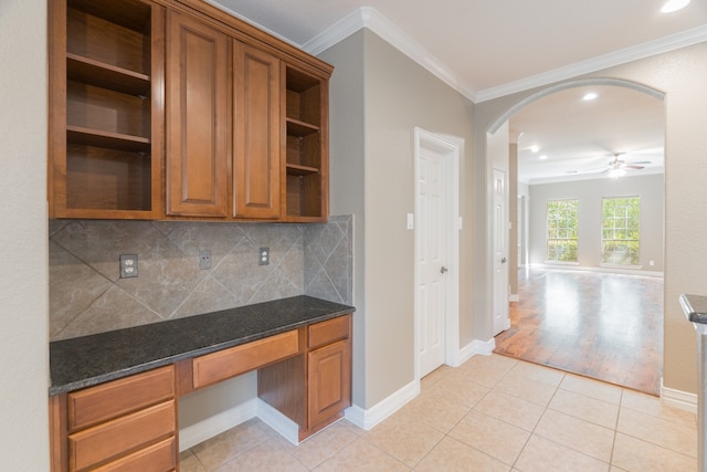 kitchen with ceiling fan, built in desk, dark stone countertops, light wood-type flooring, and crown molding