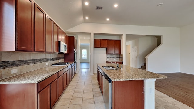 kitchen featuring appliances with stainless steel finishes, light hardwood / wood-style floors, vaulted ceiling, light stone counters, and a kitchen island with sink