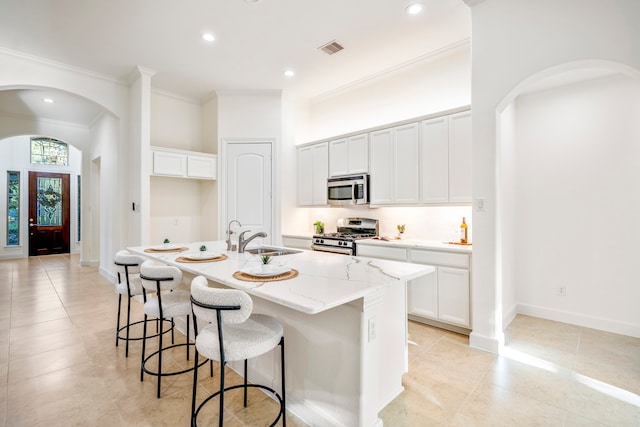 kitchen with arched walkways, a kitchen island with sink, stainless steel appliances, white cabinetry, and a sink