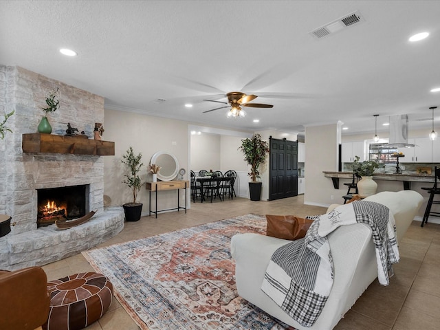 living room featuring a stone fireplace, ceiling fan, crown molding, and light tile patterned floors