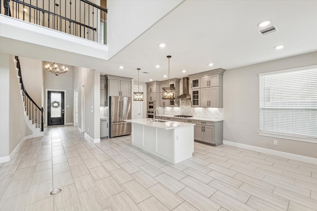 kitchen featuring tasteful backsplash, hanging light fixtures, wall chimney exhaust hood, stainless steel appliances, and a center island with sink