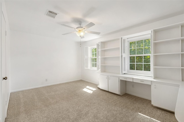 unfurnished office with built in desk, a healthy amount of sunlight, light colored carpet, and ceiling fan